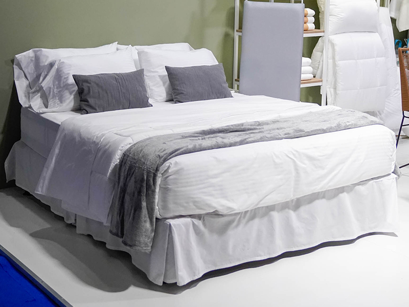 foto de Cama matrimonial con ropa de cama blanca y detalles en gris, exhibida como equipamiento hotelero en el evento Hotelga.