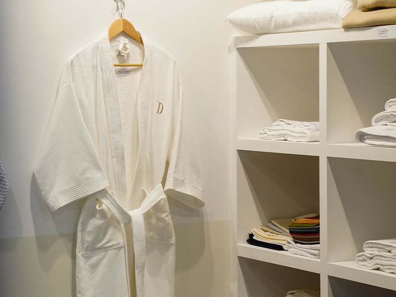 foto de Bata de baño blanca de estilo hotelero exhibida en un perchero, junto a toallas y ropa de cama en estanterías de un stand de la exposición Hotelga.