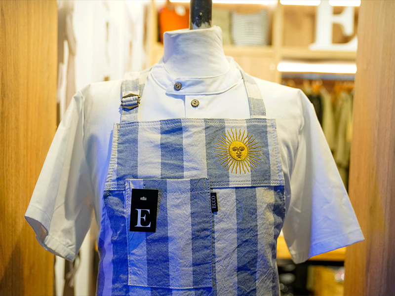 foto de Uniforme gastronómico con delantal estampado en tonos celeste y blanco, exhibido en un stand de indumentaria profesional en el evento Hotelga.