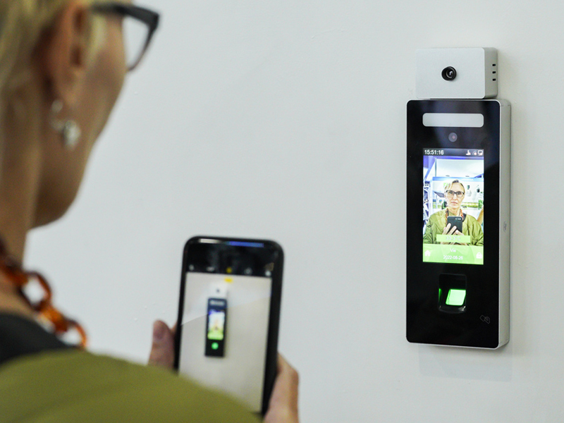 Una mujer toma una foto con su teléfono móvil de un dispositivo de reconocimiento facial montado en una pared blanca. La pantalla del dispositivo muestra la imagen de una persona.