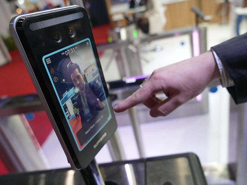 Una mano está tocando la pantalla de un dispositivo de reconocimiento facial que muestra una selfie de un hombre y la hora.
