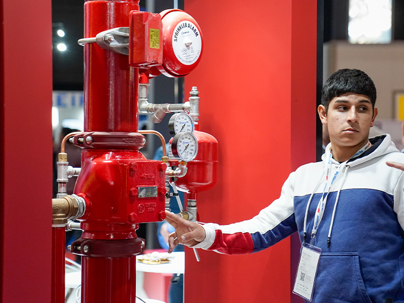 Un joven señala un sistema de tuberías rojas con manómetros y una alarma contra incendios.