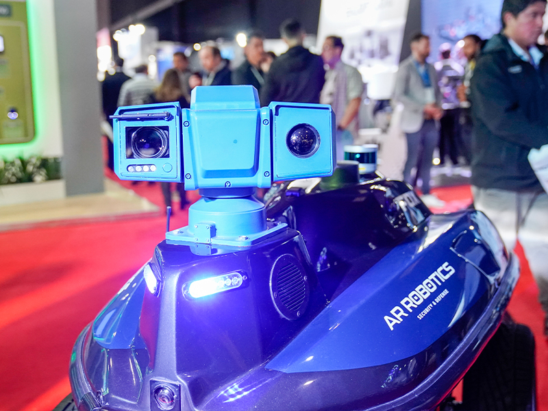 Un robot de seguridad azul con múltiples cámaras, exhibido en Intersec BA con gente en el fondo.