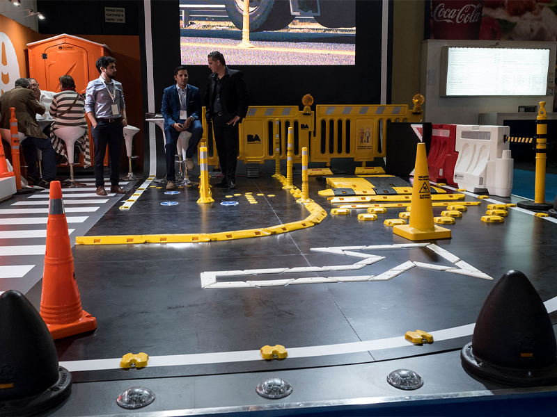 Un stand que simula una zona de construcción con conos de tráfico naranjas, barreras amarillas y señales de seguridad en el suelo. Varias personas están de pie o sentadas en el fondo.