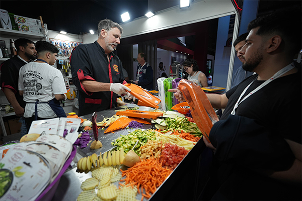 Expositor en su stand mostrando un utensilio de cocina naranja para cortar verduras a un grupo de personas. Sobre la mesa hay una variedad de verduras frescas ya cortadas en diferentes formas, como zanahorias, calabacines y patatas en espiral.