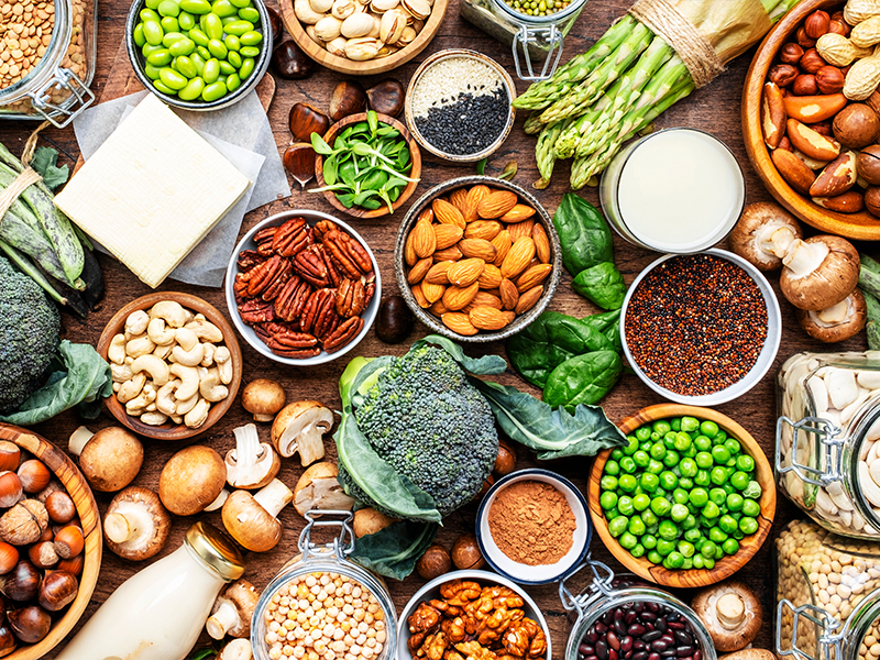 Una vista aérea de varios cuencos de madera y recipientes con una gran variedad de alimentos ricos en proteínas vegetales, como almendras, nueces, frijoles, garbanzos, tofu, brócoli, champiñones, guisantes y quinoa.
