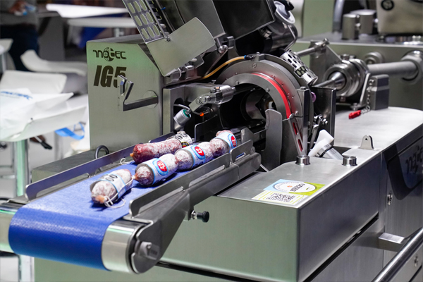 Primer plano de una máquina industrial moderna, de acero inoxidable. La máquina está en funcionamiento y se observa una cinta transportadora de color azul por donde se desplazan salchichas ya envasadas.